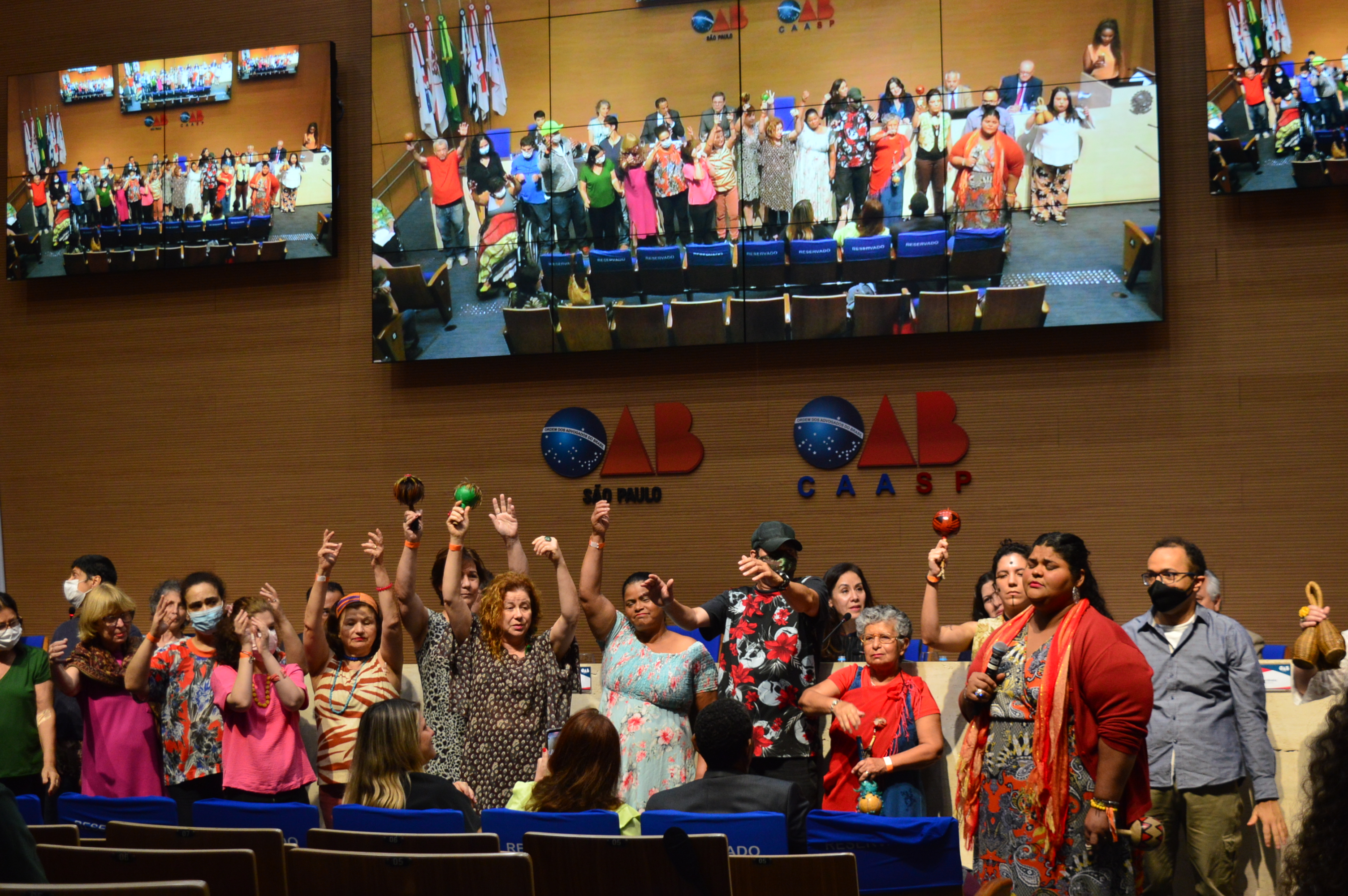 Um grupo de pessoas está em uma apresentação de canto com as mãos levantadas com chocalhos e uma delas, uma mulher vestida de vermelho está com o microfone nas mãos. Todos estão com em frente à plenária da OAB SP.