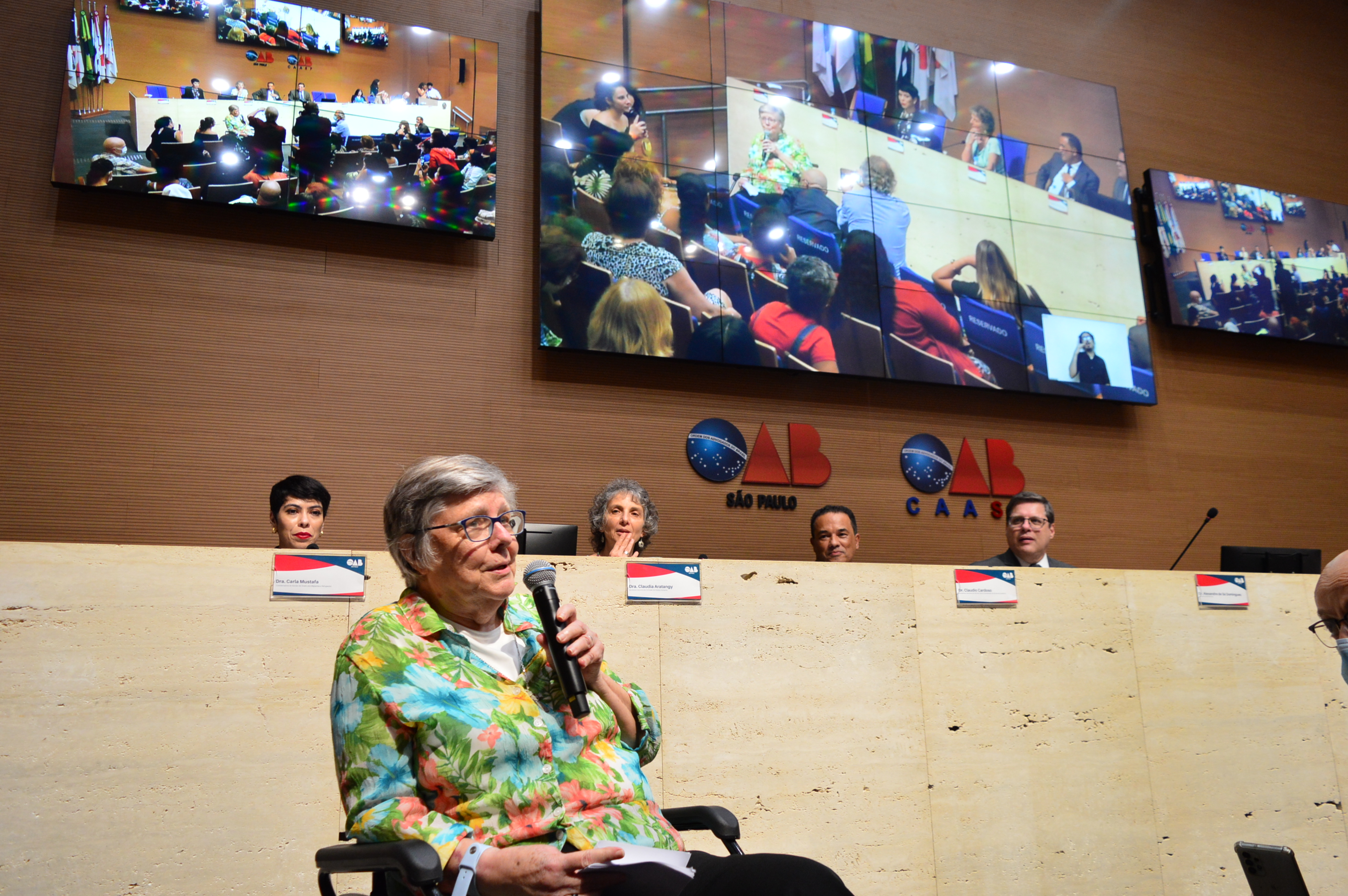 Uma senhora branca de cabelos grisalhos e óculos segurando um microfone discursa. Atrás dela, quatro pessoas sentadas numa mesa e três telões transmitem o evento.