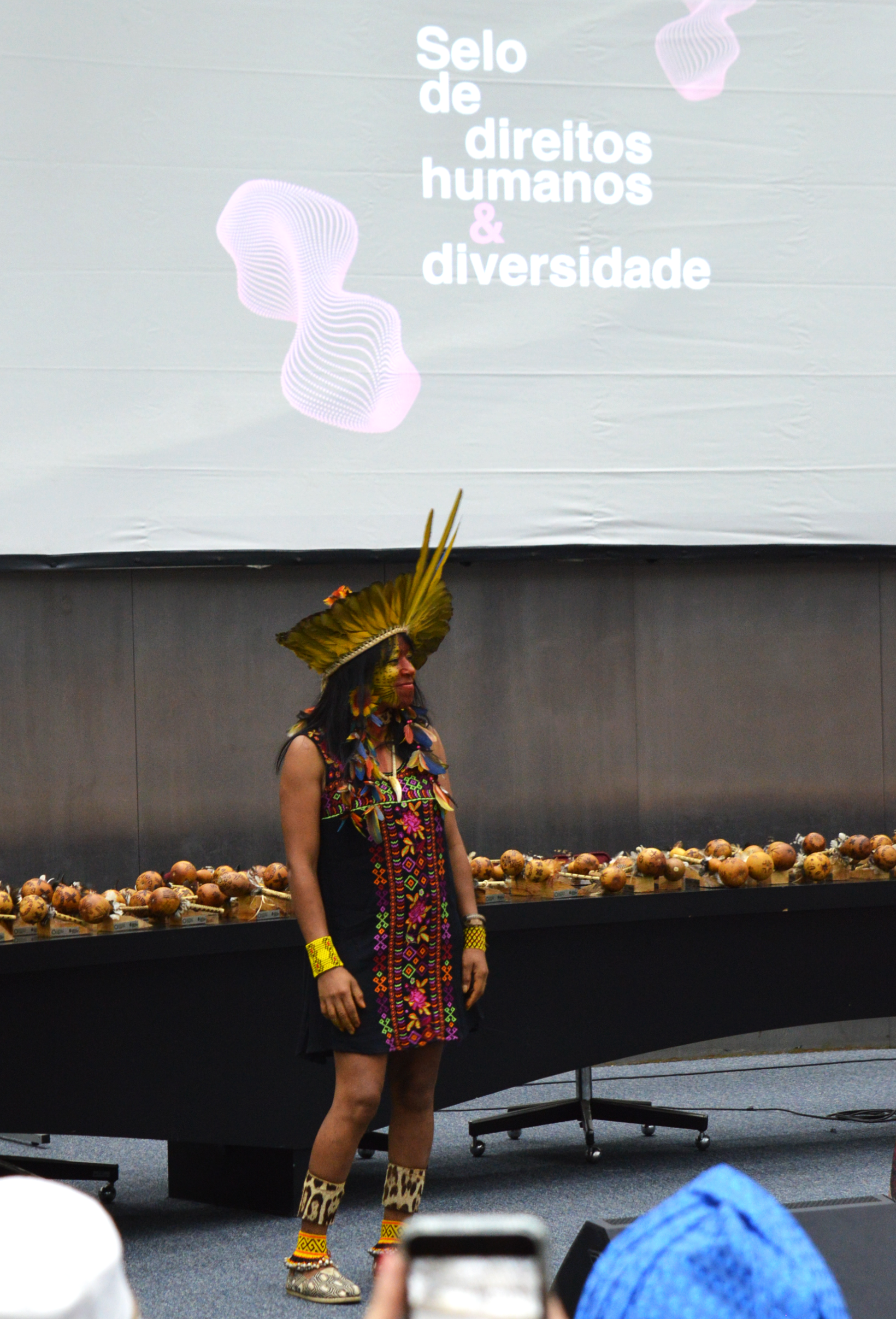A artista Tamikuã Thixi é uma mulher indígena, está vestida com uma roupa preta com detalhes floridos. Usa um cocar de penas amarelas e azuis. Ao fundo estão as estatuetas do prêmio.